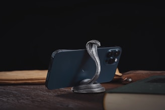 A smartphone is supported by a metallic snake-shaped stand on a wooden surface. The background is dark, and the phone is surrounded by books and papers.