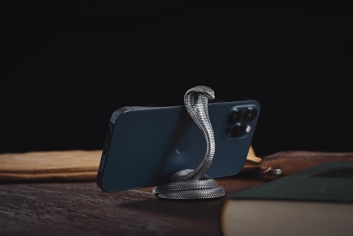 A smartphone is supported by a metallic snake-shaped stand on a wooden surface. The background is dark, and the phone is surrounded by books and papers.
