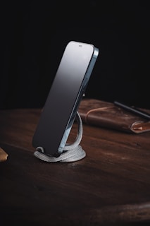 A sleek smartphone is propped up on a coiled, silver-colored metal stand placed on a wooden surface. In the background, a brown leather item, possibly a notebook or a wallet, and a pen are partially visible. The setting conveys an organized and minimalist workspace.