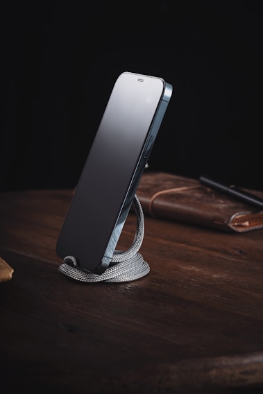 A sleek smartphone is propped up on a coiled, silver-colored metal stand placed on a wooden surface. In the background, a brown leather item, possibly a notebook or a wallet, and a pen are partially visible. The setting conveys an organized and minimalist workspace.