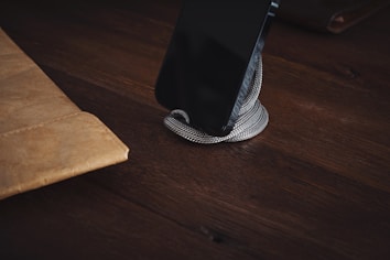 A snake is coiled around a smartphone, placed on a dark wooden surface. To the left, a brown leather folder or sleeve is partially visible. The setting is dimly lit, giving a mysterious and rustic feel.
