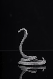 A silver metallic sculpture of a snake is coiled elegantly with its head slightly raised, set against a dark background. The intricate scales of the snake reflect the light, adding texture and depth.