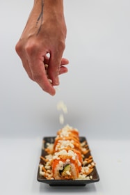 A hand with a tattoo is sprinkling white toppings onto a row of sushi rolls arranged on a black rectangular plate. The sushi is topped with white and orange-colored ingredients, creating a visually appealing arrangement.