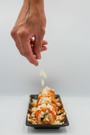 A hand with a tattoo is sprinkling white toppings onto a row of sushi rolls arranged on a black rectangular plate. The sushi is topped with white and orange-colored ingredients, creating a visually appealing arrangement.