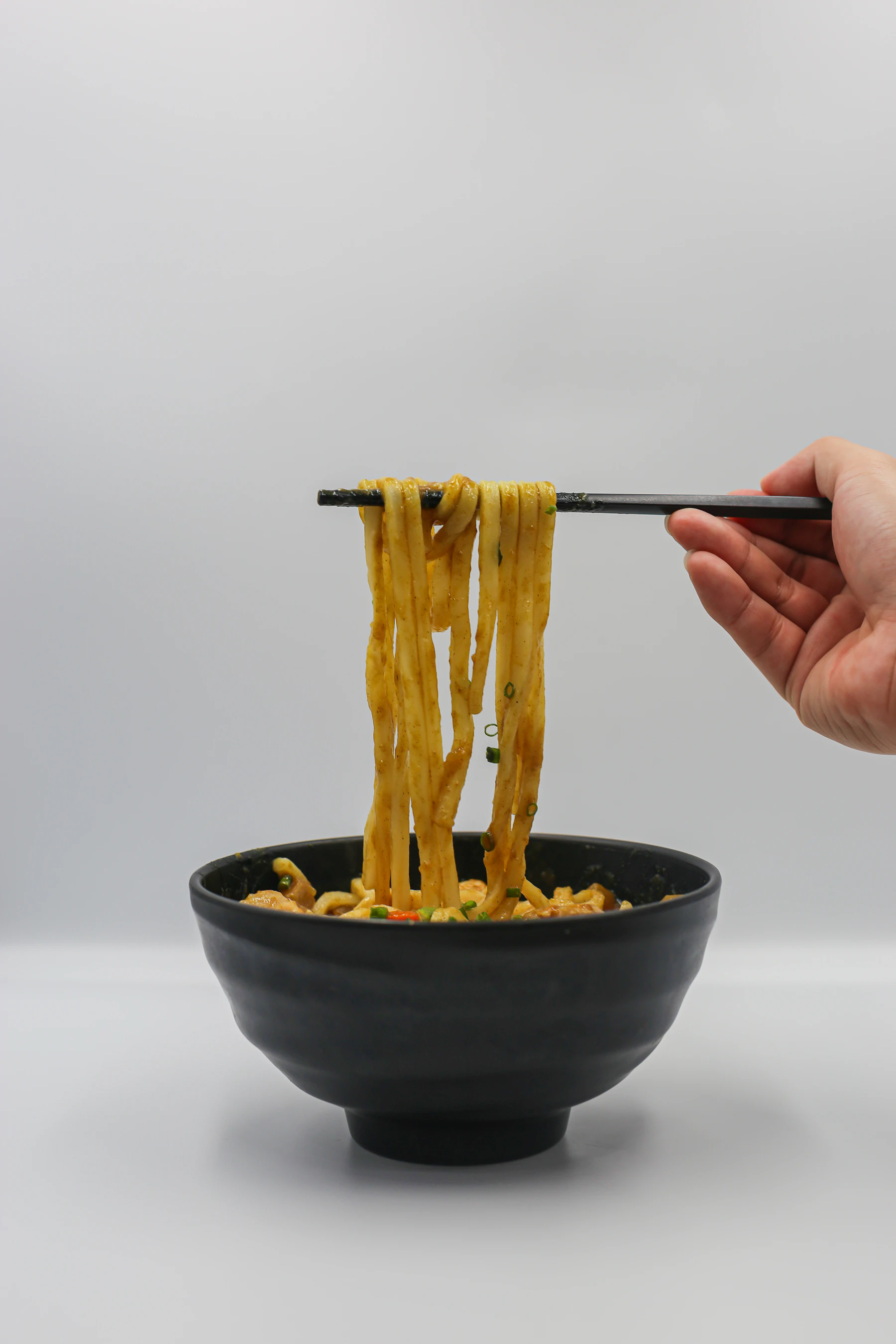 A hand lifting thick yellow ramen noodles out of a bowl with chopsticks