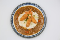 A round plate contains a serving of crispy golden-brown fried shrimp placed on a bed of white rice. The rice is surrounded by a rich, brown curry sauce with visible chunks of carrots and potatoes. The dish is garnished with finely chopped green herbs.