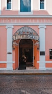 An entrance to a hotel with a pink facade and white accents. The doorway features a dome-like awning with floral designs and the words 'Hotel Merida' inscribed on it. Two lanterns flank the entrance, and the street in front is paved with patterned stone.