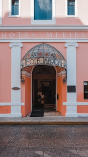 An entrance to a hotel with a pink facade and white accents. The doorway features a dome-like awning with floral designs and the words 'Hotel Merida' inscribed on it. Two lanterns flank the entrance, and the street in front is paved with patterned stone.