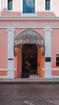 An entrance to a hotel with a pink facade and white accents. The doorway features a dome-like awning with floral designs and the words 'Hotel Merida' inscribed on it. Two lanterns flank the entrance, and the street in front is paved with patterned stone.