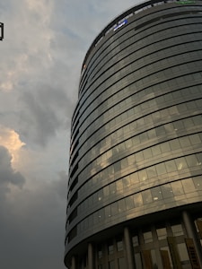 A modern cylindrical glass building with multiple floors, is seen against a cloudy sky. The building features reflective windows that capture the light and clouds. There are signs of professional services companies at the top and the bottom sections supported by columns.