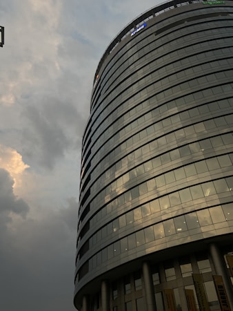 A modern cylindrical glass building with multiple floors is seen against a cloudy sky. The building features reflective windows that capture the light and clouds. There are signs of professional services companies at the top and the bottom sections supported by columns.