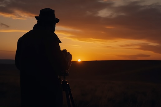 A silhouette of a person wearing a hat is captured against a dramatic sunset. The scene features a tripod and camera as the sun descends near the horizon, enveloped by a vibrant orange and darkening sky.