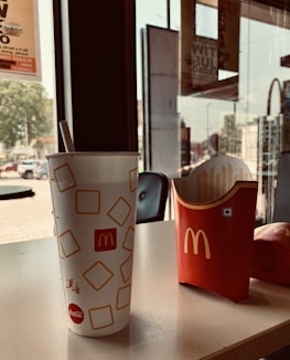A fast-food restaurant setting with a large soft drink cup and an empty french fry container, both branded with McDonald's logo, placed on a table near a window. The background includes a slight view of the outdoor environment and interior space.