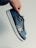 A hand is holding a sneaker with a sleek design. The shoe features a combination of black leather and blue accents, with a black Nike swoosh. The sole is white with blue detailing on the bottom.