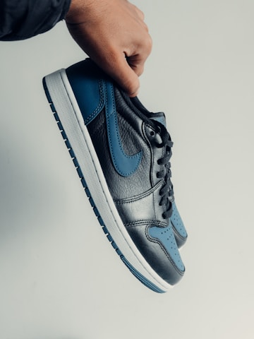 A hand is holding a sneaker with a sleek design. The shoe features a combination of black leather and blue accents, with a black Nike swoosh. The sole is white with blue detailing on the bottom.