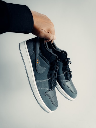 A hand is holding a pair of dark-colored athletic shoes. The shoes have a combination of leather and mesh materials with a swoosh logo on the side. They feature black laces and white soles. The hand appears to be wearing a black sleeve.