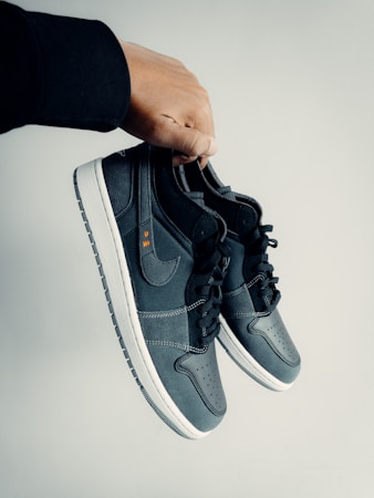 A hand is holding a pair of dark-colored athletic shoes. The shoes have a combination of leather and mesh materials with a swoosh logo on the side. They feature black laces and white soles. The hand appears to be wearing a black sleeve.