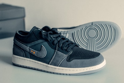 A pair of stylish sneakers with a sleek design and a mix of dark and light gray colors. The shoes display a prominent logo on the side with a textured pattern on the sole. The laces are neatly tied, and the sneakers are placed on a smooth surface with a box in the background.