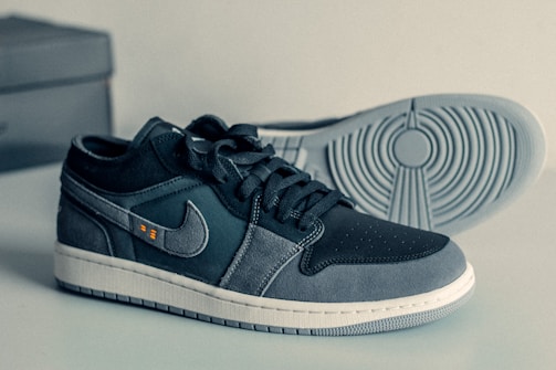 A pair of stylish sneakers with a sleek design and a mix of dark and light gray colors. The shoes display a prominent logo on the side with a textured pattern on the sole. The laces are neatly tied, and the sneakers are placed on a smooth surface with a box in the background.