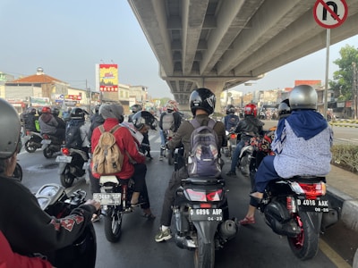 A group of people on motorcycles are waiting at a junction under an overpass. Most of them are wearing helmets and backpacks. There are various buildings and signs in the background, one depicting a bright sunburst pattern. A no U-turn sign is visible overhead.