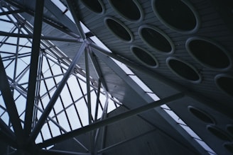 A complex architectural structure featuring intersecting metal beams set against a backdrop of a glass ceiling. The ceiling incorporates multiple circular elements, likely functioning as skylights or design features. The perspective provides a dynamic view of the geometry involved in the construction.