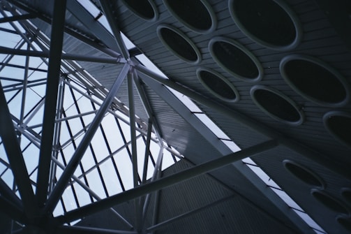 A complex architectural structure featuring intersecting metal beams set against a backdrop of a glass ceiling. The ceiling incorporates multiple circular elements, likely functioning as skylights or design features. The perspective provides a dynamic view of the geometry involved in the construction.