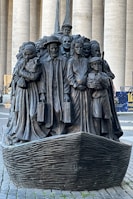A bronze sculpture depicting a diverse group of people standing closely together on a boat, with expressions and attire representing various cultures and eras. The figures include men, women, and children, some holding luggage, symbolizing themes of migration and unity.