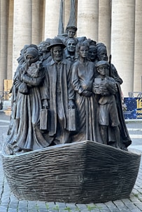 A bronze sculpture depicting a diverse group of people standing closely together on a boat, with expressions and attire representing various cultures and eras. The figures include men, women, and children, some holding luggage, symbolizing themes of migration and unity.