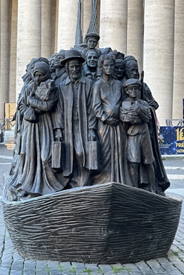A bronze sculpture depicting a diverse group of people standing closely together on a boat, with expressions and attire representing various cultures and eras. The figures include men, women, and children, some holding luggage, symbolizing themes of migration and unity.