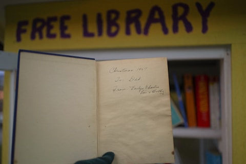 An open book is held up in front of a small structure or box labeled 'Free Library' in purple letters on a yellow background. The book has an inscription reading 'Christmas 1997. To Dad. From Evelyn, Charlie, Kris & Betty'. Inside the library, a few books with colorful spines are partially visible.