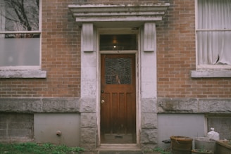 A weathered wooden door with a decorative glass panel is set in a brick building. The facade includes stone elements around the entrance. Two windows with partially drawn curtains flank the door, and there are some metal and plastic containers in the corner near an overgrown patch of grass.