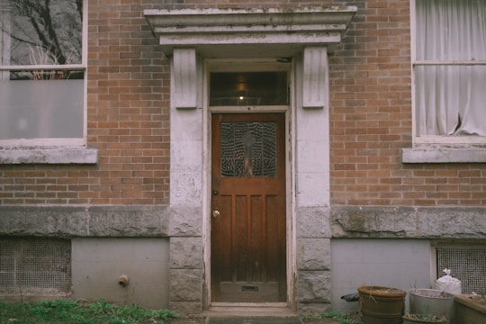 A weathered wooden door with a decorative glass panel is set in a brick building. The facade includes stone elements around the entrance. Two windows with partially drawn curtains flank the door, and there are some metal and plastic containers in the corner near an overgrown patch of grass.