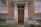A weathered wooden door with a decorative glass panel is set in a brick building. The facade includes stone elements around the entrance. Two windows with partially drawn curtains flank the door, and there are some metal and plastic containers in the corner near an overgrown patch of grass.
