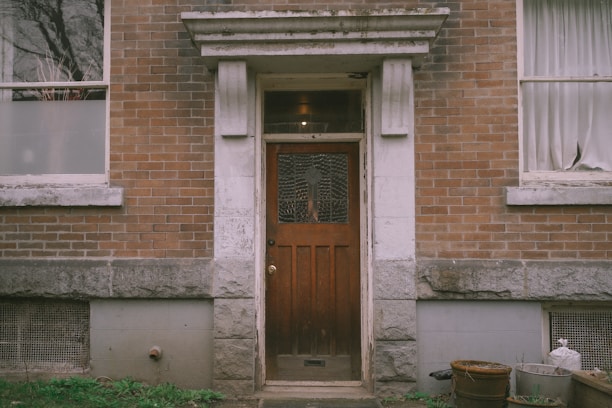 A weathered wooden door with a decorative glass panel is set in a brick building. The facade includes stone elements around the entrance. Two windows with partially drawn curtains flank the door, and there are some metal and plastic containers in the corner near an overgrown patch of grass.