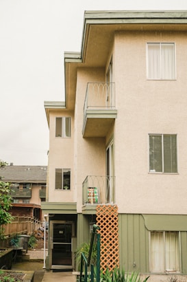 A multi-story residential building exterior with light beige walls and green trim. The building features balconies with metal railings and several windows. In the foreground, there is a green railing beside a staircase that leads to an entrance door marked with the number 187. A lattice panel is attached to the side of the building. To the left, a wooden fence separates the property from a neighboring building.