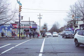 A busy street intersection featuring a Hasty Market on the corner with several people gathered outside. Multiple vehicles are visible on the road. The street is lined with bare trees and buildings, and murals decorate the walls. The sky is overcast, creating a muted ambience.