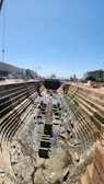 A large, empty dry dock with stepped sides and a central rail track. It is surrounded by industrial buildings, cranes, and dockside equipment under a clear blue sky. The dry dock has some remnants of water and scattered debris at the bottom.