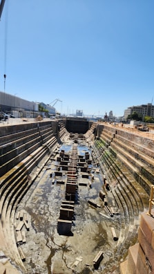 A large, empty dry dock with stepped sides and a central rail track. It is surrounded by industrial buildings, cranes, and dockside equipment under a clear blue sky. The dry dock has some remnants of water and scattered debris at the bottom.