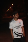 A young man wearing glasses and a sports jersey is standing outdoors at night. The street lights in the background create a line of small glowing orbs, illuminating the path in the distance. The jersey reads 'M.Bin Qasim House' and features a logo on the left side of his chest.