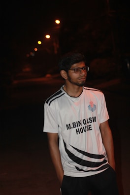 A young man wearing glasses and a sports jersey is standing outdoors at night. The street lights in the background create a line of small glowing orbs, illuminating the path in the distance. The jersey reads 'M.Bin Qasim House' and features a logo on the left side of his chest.