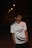 A young man wearing glasses and a sports jersey is standing outdoors at night. The street lights in the background create a line of small glowing orbs, illuminating the path in the distance. The jersey reads 'M.Bin Qasim House' and features a logo on the left side of his chest.