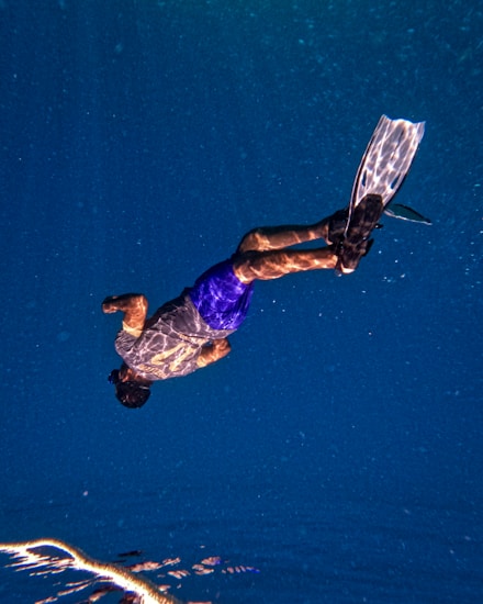 A person is swimming underwater, wearing a pair of blue shorts and using black fins. The deep blue water surrounds them, with light patterns dancing across their skin and equipment due to the refraction from the water surface above.