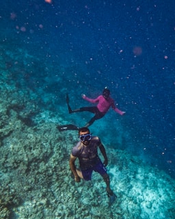Two individuals are engaged in snorkeling underwater. The clear blue water surrounds them, providing a view of the coral reef below. One person is wearing a short-sleeved shirt and swim trunks, while the other wears a long-sleeved pink shirt. Both have diving masks and are equipped with fins.