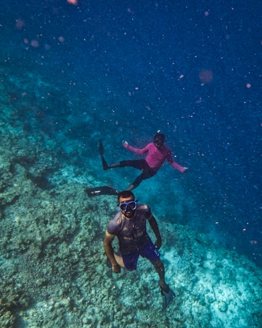 Two individuals are engaged in snorkeling underwater. The clear blue water surrounds them, providing a view of the coral reef below. One person is wearing a short-sleeved shirt and swim trunks, while the other wears a long-sleeved pink shirt. Both have diving masks and are equipped with fins.