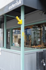 The image captures the exterior of a shop with a partly visible awning under the sign 'asterisk'. The storefront includes a window display showing tools or crafted items. The design features light blue-green wooden framing and grey panels, with a yellow sign hanging outwards.