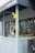 The image captures the exterior of a shop with a partly visible awning under the sign 'asterisk'. The storefront includes a window display showing tools or crafted items. The design features light blue-green wooden framing and grey panels, with a yellow sign hanging outwards.