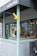The image captures the exterior of a shop with a partly visible awning under the sign 'asterisk'. The storefront includes a window display showing tools or crafted items. The design features light blue-green wooden framing and grey panels, with a yellow sign hanging outwards.