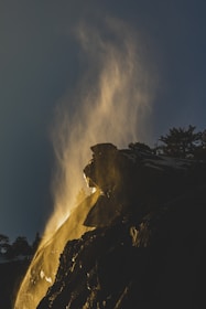 A dramatic scene featuring mist or steam rising from a rocky cliff, illuminated by warm, golden light, against a dark sky. Silhouetted trees are visible at the edge of the cliff.