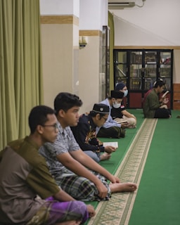 Several people are sitting on a green carpeted floor in a room that appears to be a mosque or a prayer room. Some are reading from books, while others sit quietly. The room has a bookshelves filled with books and a curtain on one side.