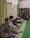 Several people are sitting on a green carpeted floor in a room that appears to be a mosque or a prayer room. Some are reading from books, while others sit quietly. The room has a bookshelves filled with books and a curtain on one side.